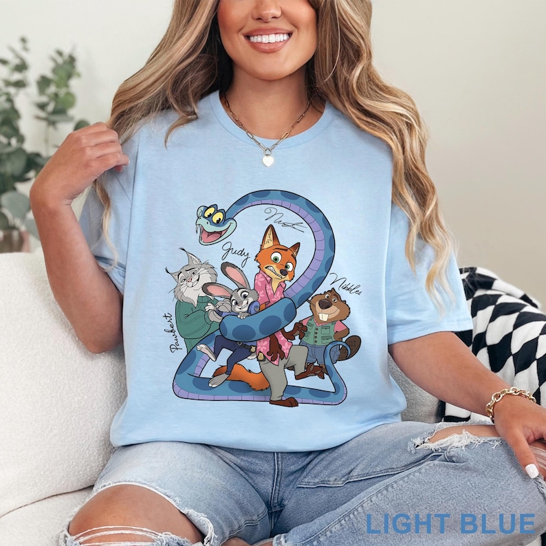 Puede incluir: Una camiseta azul claro con un estampado gr&aacute;fico de personajes de dibujos animados, incluyendo un zorro, un conejo, un lince, un castor y una serpiente. El texto "Judy", "Nick", "Nibbles" y "Pawbert" est&aacute; impreso en negro cerca de ellos.