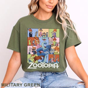 May include: An olive green t-shirt featuring a large rectangular graphic print with multiple scenes of animated characters from Zootopia. The design includes a central blue snake surrounded by various character panels and the white Zootopia logo text.