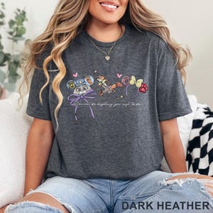 May include: A dark heather gray short-sleeve t-shirt featuring a graphic print of a rabbit character with police ears, a fox character, and a donut-themed ear headband. The text reads, You can be anything you wish to be, with small pink hearts.