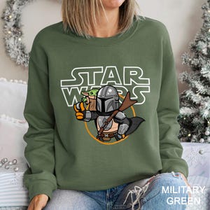 May include: Military green sweatshirt featuring a cartoon illustration of The Mandalorian and Grogu from Star Wars. The Star Wars logo is printed above the characters. The sweatshirt is a crew neck style.