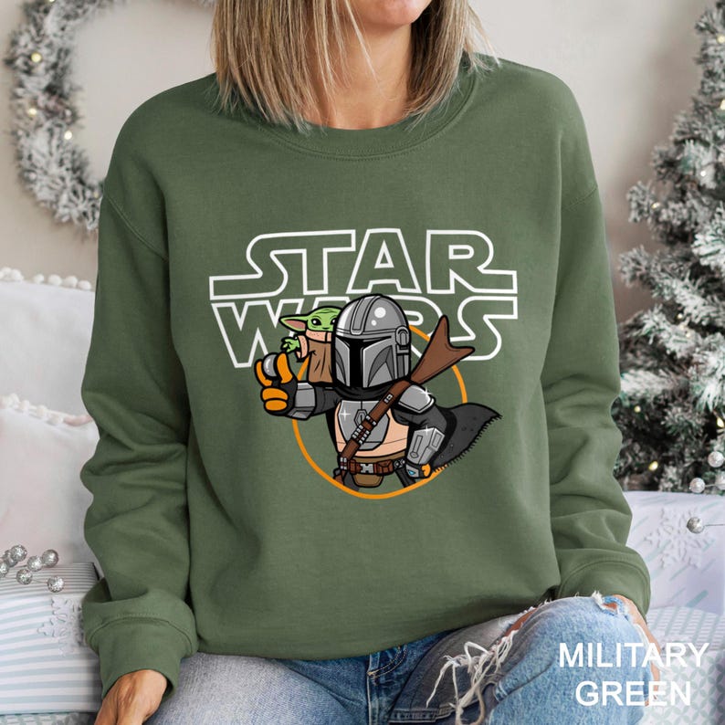 May include: Military green sweatshirt featuring a cartoon illustration of The Mandalorian and Grogu from Star Wars. The Star Wars logo is printed above the characters. The sweatshirt is a crew neck style.
