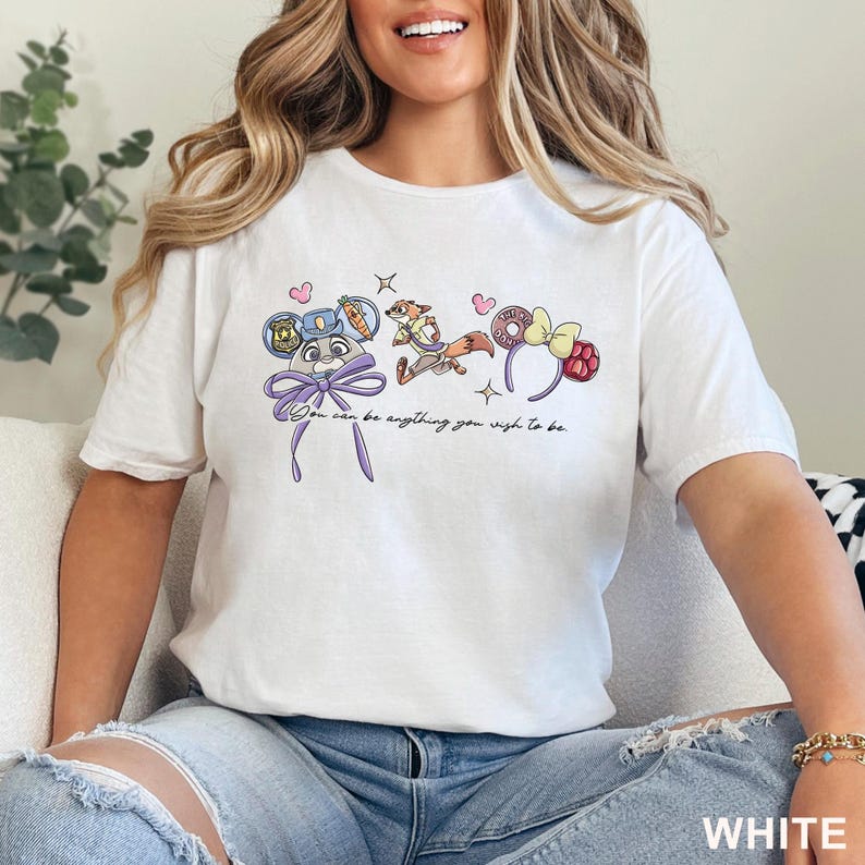 May include: A white short-sleeve t-shirt featuring a graphic print of a rabbit character with blue ears, a fox character, and a headband with a yellow bow. The text reads, "You can be anything you wish to be." The design includes small pink hearts and stars.