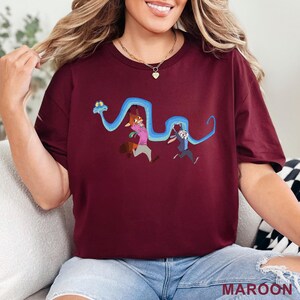 May include: A maroon short-sleeved t-shirt featuring a graphic print of a long blue snake winding around a cartoon fox and a cartoon rabbit. The fox wears a pink shirt and the rabbit wears a blue vest. The design is centered on the chest area.