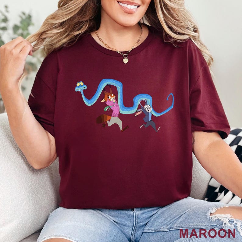 May include: A maroon short-sleeved t-shirt featuring a graphic print of a long blue snake winding around a cartoon fox and a cartoon rabbit. The fox wears a pink shirt and the rabbit wears a blue vest. The design is centered on the chest area.