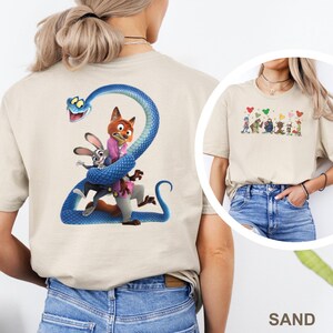 K&ouml;nnte beinhalten: Sandfarbenes T-Shirt mit einer farbenfrohen Grafik von Charakteren aus einem beliebten Animationsfilm. Auf der R&uuml;ckseite des Shirts ist eine blaue Schlange zu sehen, die sich um einen Fuchs, ein Kaninchen und eine Figur windet. Die Vorderseite hat eine kleinere Grafik von Charakteren und Herzen.