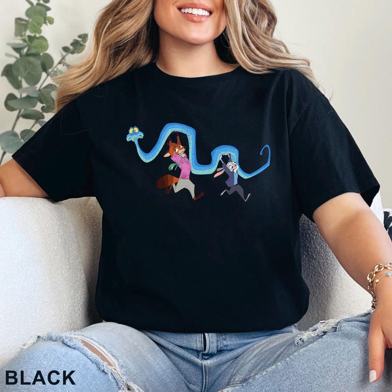 May include: A black short-sleeved t-shirt featuring a graphic print of a long, winding blue snake held by a cartoon fox wearing a pink shirt and a cartoon rabbit wearing a blue shirt. The characters are depicted in a running pose with the snake.
