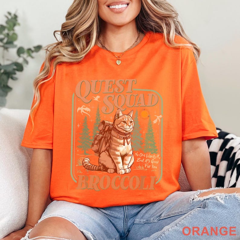 May include: Orange t-shirt with a graphic of a cat wearing a backpack, set in a forest scene with the words "QUEST SQUAD" and "BROCCOLI". The shirt also has the text "No One Wants It, But It's Good for You".