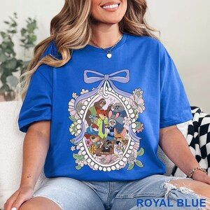 Pu&ograve; includere: T-shirt blu reale con un'illustrazione di personaggi di animali dei cartoni animati all'interno di una cornice ovale, decorata con fiori e un fiocco. Il testo "ROYAL BLUE" &egrave; in basso. La maglietta &egrave; di colore blu reale.