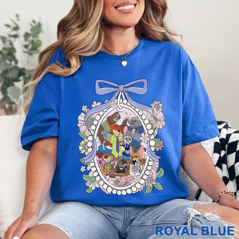 Pu&ograve; includere: T-shirt blu reale con un'illustrazione di personaggi di animali dei cartoni animati all'interno di una cornice ovale, decorata con fiori e un fiocco. Il testo "ROYAL BLUE" &egrave; in basso. La maglietta &egrave; di colore blu reale.