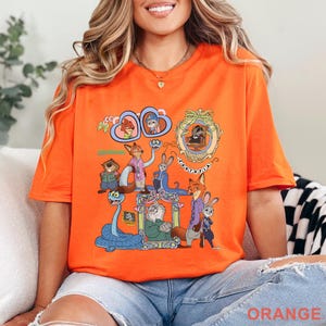 May include: Orange t-shirt featuring a colorful graphic of characters from the animated movie Zootopia. The design includes various characters in ornate frames and the word "Zootopia". The shirt is made of a soft material and has short sleeves.