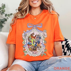 Pu&ograve; includere: T-shirt arancione brillante con un'illustrazione di cartoni animati di personaggi di animali in una cornice ovale floreale, sormontata da un fiocco viola. La parola "ORANGE" &egrave; stampata in basso sulla maglietta.