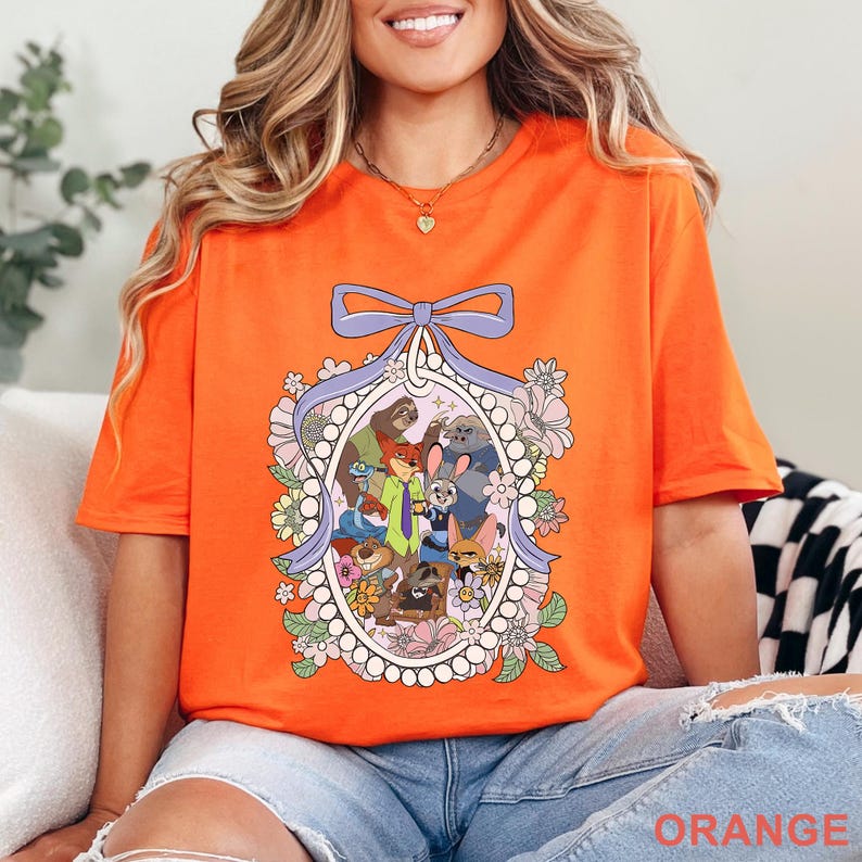 Pu&ograve; includere: T-shirt arancione brillante con un'illustrazione di cartoni animati di personaggi di animali in una cornice ovale floreale, sormontata da un fiocco viola. La parola "ORANGE" &egrave; stampata in basso sulla maglietta.