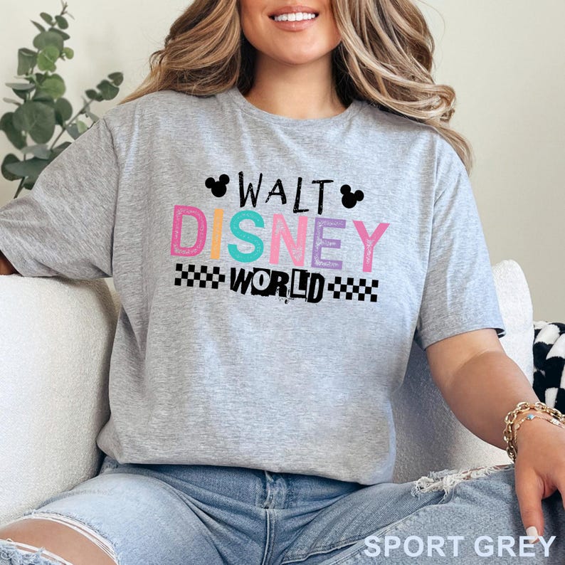 Pu&ograve; includere: Una t-shirt grigio melange a maniche corte con la scritta WALT DISNEY WORLD in un carattere multicolore dall'aspetto usurato. Il design include due sagome nere di orecchie di Topolino e un motivo a scacchi bianco e nero sotto la parola WORLD.