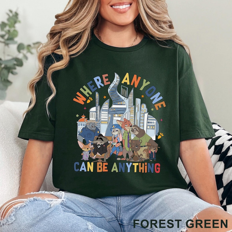 May include: Forest green t-shirt featuring a colorful graphic with the text "WHERE ANYONE CAN BE ANYTHING" above a cartoon cityscape and animal characters. The shirt is a crew neck style.