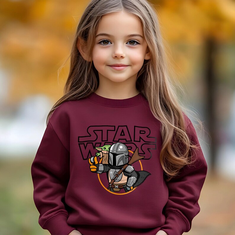 May include: Burgundy sweatshirt featuring a Star Wars graphic. The graphic includes the Star Wars logo and an illustration of The Mandalorian and Grogu. The sweatshirt is worn by a person with long brown hair.
