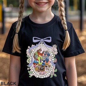 Pu&ograve; includere: T-shirt nera con una grafica a cartoni animati colorata di vari personaggi di animali all'interno di una cornice ovale floreale, con un fiocco viola chiaro in alto. La parola "BLACK" &egrave; stampata in basso sulla maglietta.