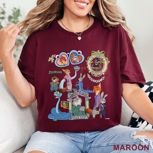 May include: Maroon t-shirt featuring a colorful graphic of characters from the animated film Zootopia. The design includes various characters in ornate frames and the film's title. The shirt is a solid maroon color.