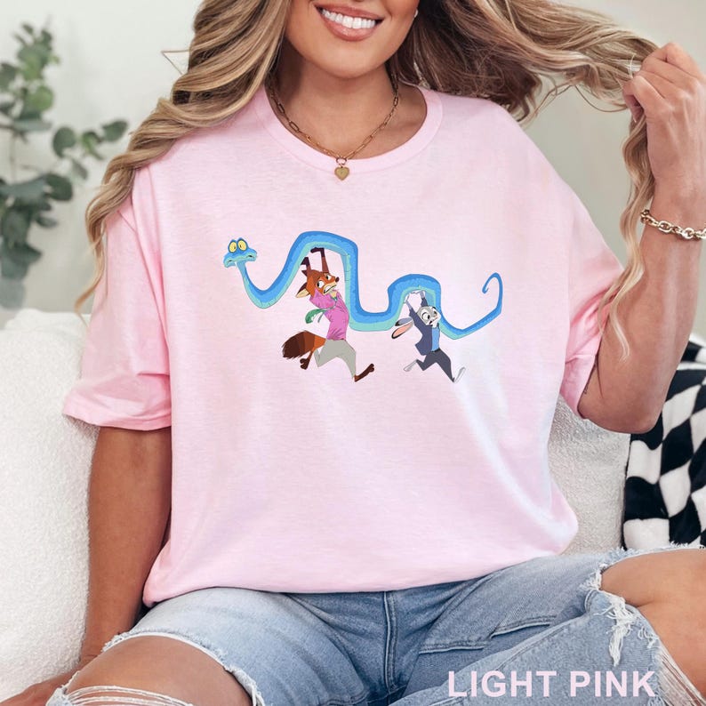 May include: A light pink t-shirt featuring a graphic print of a long blue snake winding around a cartoon fox wearing a pink shirt and gray pants, and a cartoon rabbit wearing a blue suit. The design shows the characters in motion.