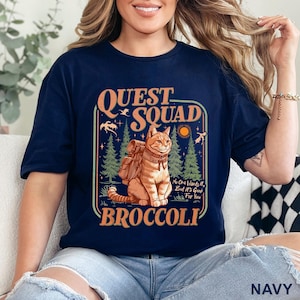 May include: Navy blue t-shirt with a graphic featuring a cat wearing a backpack, the words "Quest Squad Broccoli", and the phrase "No One Wants It, But It's Good For You". The design includes trees, a sun, and birds.