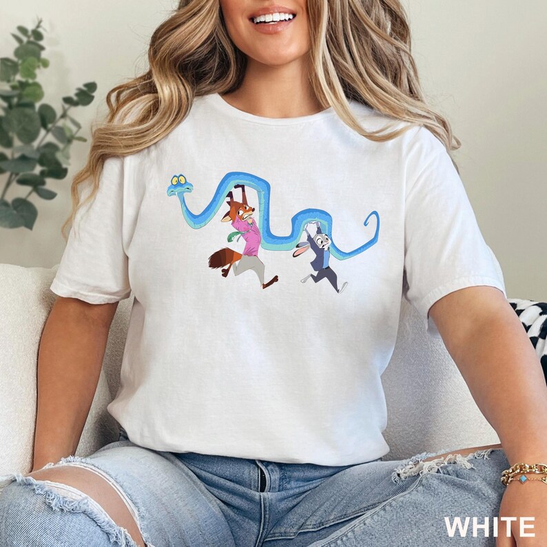 May include: A white short-sleeve t-shirt featuring a graphic print of a cartoon fox and a cartoon rabbit hanging from a long, winding blue snake. The fox wears a pink shirt and the rabbit wears a blue jacket. The design is centered on the chest.