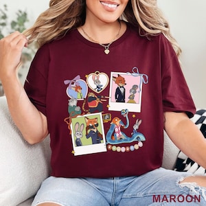 May include: A maroon short-sleeved t-shirt featuring a graphic print of Zootopia characters including Nick Wilde and Judy Hopps. The design includes multiple polaroid-style frames, heart-shaped borders, and a blue snake, with the text Zootopia.