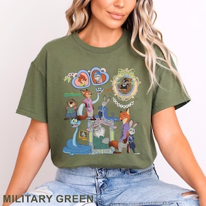 May include: Military green t-shirt featuring a colorful graphic of characters from the animated movie Zootopia. The design includes Nick Wilde, Judy Hopps, and other characters in a cartoon style. The shirt has a crew neck and short sleeves.