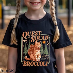 May include: Black t-shirt with a graphic featuring a cat wearing a backpack, the words "QUEST SQUAD BROCCOLI", and the phrase "No One Wants It, But It's Good For You". The design includes trees, a sun, and bats.