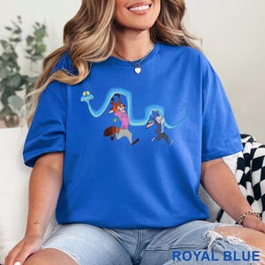 May include: A royal blue short-sleeve t-shirt featuring a graphic print of a fox and a rabbit running while being chased by a long, winding blue snake. The fox wears a pink shirt and gray pants, while the rabbit wears a blue blazer and gray pants.
