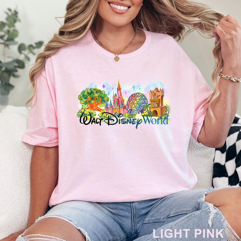 May include: A light pink short-sleeve t-shirt featuring a colorful graphic print of the Walt Disney World theme park landmarks, including a large tree, a pink castle, a geodesic dome, and a tower, with the text Walt Disney World in blue.