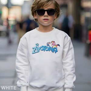 Peut inclure: Sweat-shirt blanc avec le mot "Zootopia" en bleu et des personnages de dessins anim&eacute;s. Le sweat-shirt est de style col rond. Le mot "WHITE" est imprim&eacute; en bas de l'image.