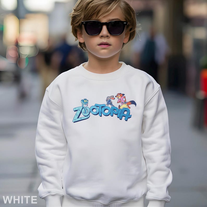 Peut inclure: Sweat-shirt blanc avec le mot "Zootopia" en bleu et des personnages de dessins anim&eacute;s. Le sweat-shirt est de style col rond. Le mot "WHITE" est imprim&eacute; en bas de l'image.