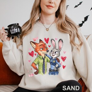 May include: Sand-coloured sweatshirt with a watercolour illustration of Nick Wilde and Judy Hopps from Zootopia, surrounded by pink hearts. The characters are embracing. The sweatshirt has a crew neck and long sleeves.