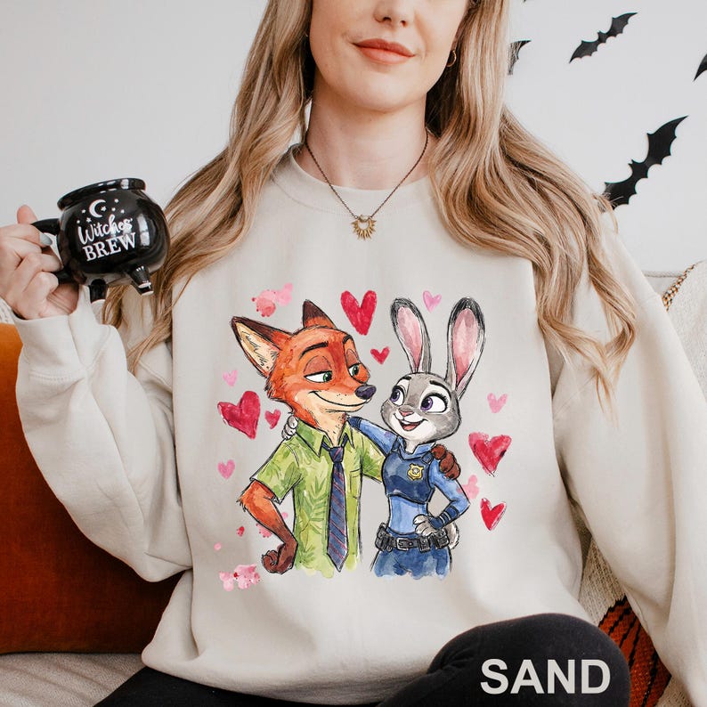 May include: Sand-coloured sweatshirt with a watercolour illustration of Nick Wilde and Judy Hopps from Zootopia, surrounded by pink hearts. The characters are embracing. The sweatshirt has a crew neck and long sleeves.