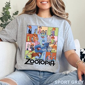 May include: A heather grey short-sleeved t-shirt featuring a graphic print of various scenes from the film Zootopia, including characters Nick Wilde and Judy Hopps. The word Zootopia is printed in bold black lettering at the bottom of the design.