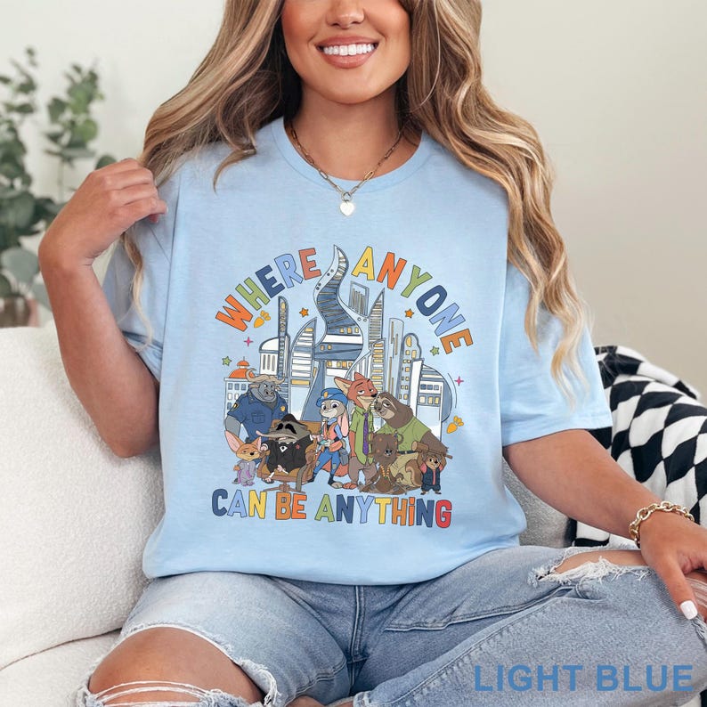 May include: Light blue t-shirt featuring a colorful graphic of cartoon characters and a cityscape. The text "WHERE ANYONE CAN BE ANYTHING" is displayed in a playful font. The shirt is made of soft material.