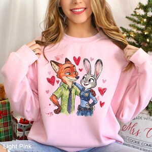 May include: Pale pink sweatshirt with a watercolour illustration of Nick Wilde and Judy Hopps from Zootopia, surrounded by red hearts. Nick is a fox in a green shirt and tie, and Judy is a rabbit in a police uniform. The text "Light Pink" is at the bottom.