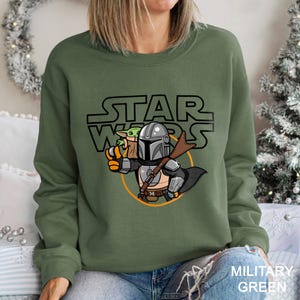 May include: Military green sweatshirt featuring a cartoon illustration of The Mandalorian holding Grogu, with the Star Wars logo above. The design includes a blaster and a circular orange outline. The text "MILITARY GREEN" is at the bottom.