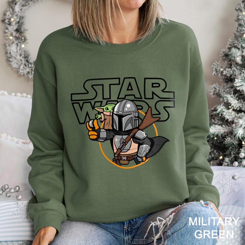 May include: Military green sweatshirt featuring a cartoon illustration of The Mandalorian holding Grogu, with the Star Wars logo above. The design includes a blaster and a circular orange outline. The text "MILITARY GREEN" is at the bottom.