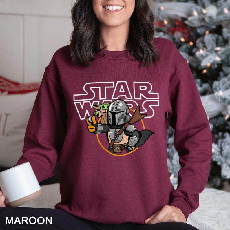 May include: Maroon sweatshirt featuring a Star Wars graphic. The design includes the text "STAR WARS" above an illustration of The Mandalorian and Grogu. The sweatshirt is a solid maroon color.