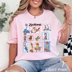 K&ouml;nnte beinhalten: Ein hellrosa T-Shirt mit einem Grafikdruck verschiedener Zootopia-Figuren in einzelnen Rahmen, darunter Judy Hopps und Nick Wilde. Der Text Zootopia ist in Schwarz oben mittig auf dem Grafikdesign auf der Vorderseite des T-Shirts gedruckt.