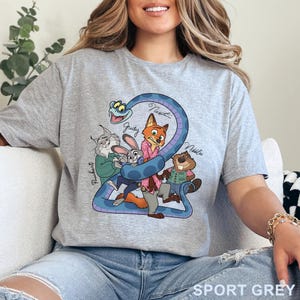 Puede incluir: Una camiseta gris claro con un estampado gr&aacute;fico de personajes de dibujos animados, incluyendo un zorro, un conejo, un lince, un castor y una serpiente azul. Los nombres Pawbert, Judy, Nick y Nibbles est&aacute;n impresos en cursiva junto a ellos.
