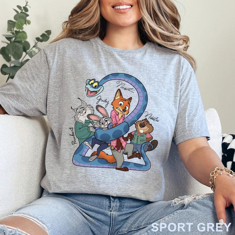 Puede incluir: Una camiseta gris claro con un estampado gr&aacute;fico de personajes de dibujos animados, incluyendo un zorro, un conejo, un lince, un castor y una serpiente azul. Los nombres Pawbert, Judy, Nick y Nibbles est&aacute;n impresos en cursiva junto a ellos.