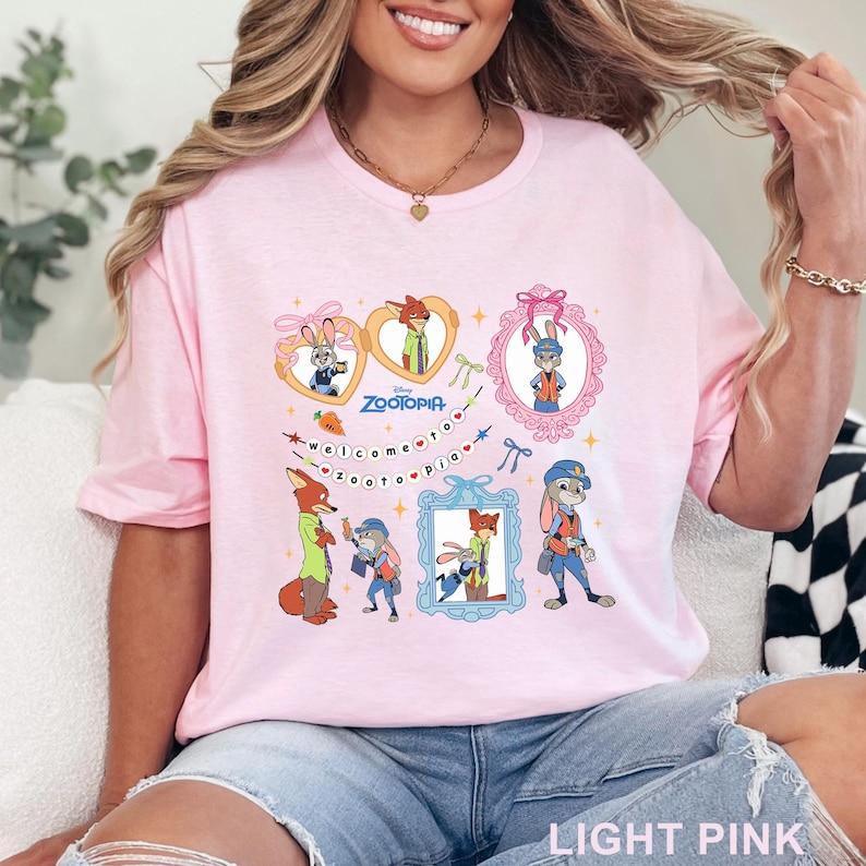 Pode incluir: T-shirt rosa claro com personagens de desenhos animados do filme Zootopia. O design inclui imagens emolduradas dos personagens, o t&iacute;tulo do filme e a frase "Bem-vindo a Zootopia". A camisa &eacute; feita de um material macio.