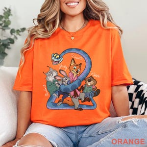 Puede incluir: Una camiseta de manga corta de color naranja brillante con un estampado gr&aacute;fico de personajes de dibujos animados, incluyendo una serpiente, un conejo, un zorro, un lince y un castor. El dise&ntilde;o incluye los nombres Pawbert, Judy, Nick y Nibbles en blanco.