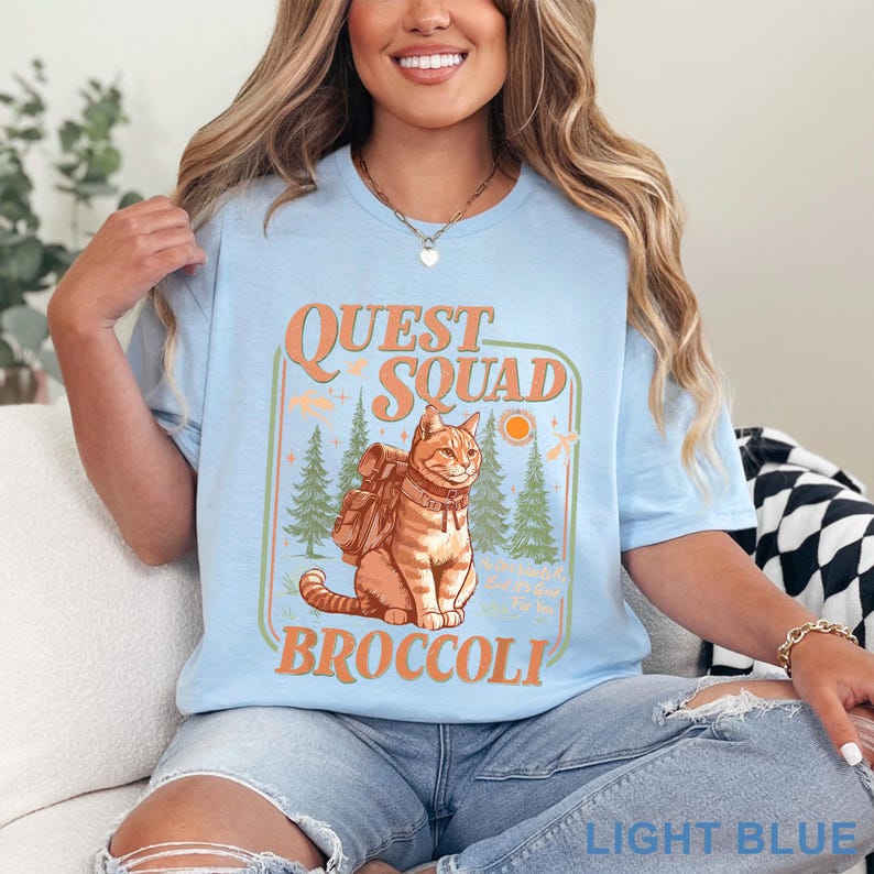 May include: Light blue t-shirt with a graphic featuring a cat wearing a backpack, the words "Quest Squad Broccoli", and a forest scene. The text "Light Blue" is at the bottom of the shirt.