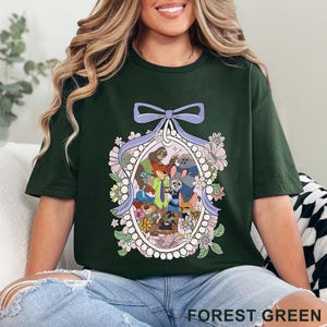 Pu&ograve; includere: T-shirt verde foresta con un'illustrazione di cartoni animati di personaggi di animali all'interno di una cornice ovale, circondata da fiori e un fiocco. Il testo "FOREST GREEN" &egrave; stampato in basso sulla maglietta.