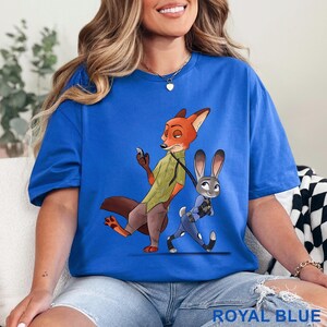 May include: Royal blue t-shirt featuring a cartoon illustration of a fox and a rabbit. The fox is wearing a green vest and brown pants, while the rabbit is in a blue uniform. The text "ROYAL BLUE" is at the bottom of the shirt.