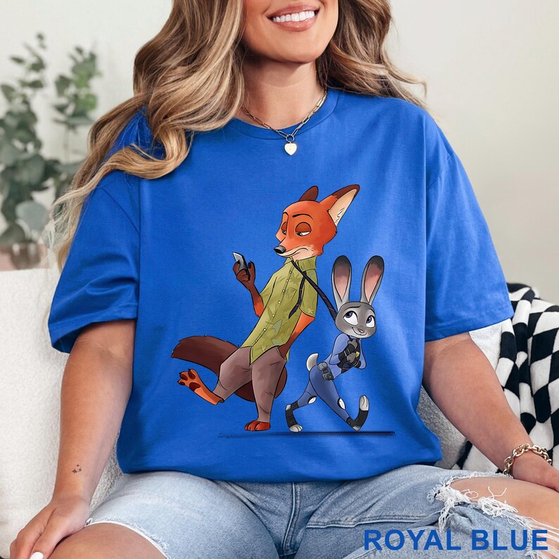 May include: Royal blue t-shirt featuring a cartoon illustration of a fox and a rabbit. The fox is wearing a green vest and brown pants, while the rabbit is in a blue uniform. The text "ROYAL BLUE" is at the bottom of the shirt.