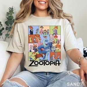 May include: A sand-coloured t-shirt featuring a grid of colourful illustrations of characters from the movie Zootopia, including a large blue snake in the centre. The word Zootopia is printed in black text at the bottom of the graphic design.