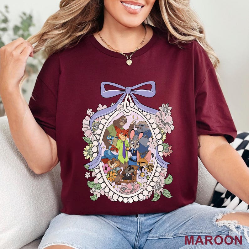 Pu&ograve; includere: T-shirt bordeaux con un'illustrazione colorata di personaggi di animali animati all'interno di una cornice floreale. Un fiocco viola in alto e la scritta "MAROON" in basso. La maglietta &egrave; a girocollo.